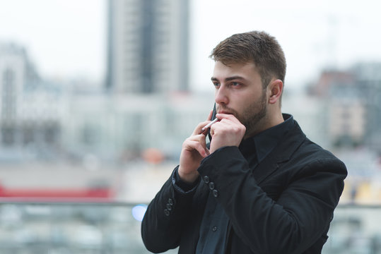 A businessman smokes a cigarette and speaks by telephone on the background of a city landscape.