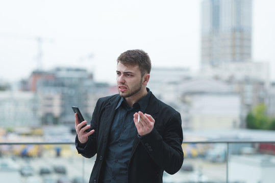 An emotional businessman with a telephone in his hands smokes a cigarette and is angry with the background of a city-blurred landscape. An angry man calms himself with cigarettes.