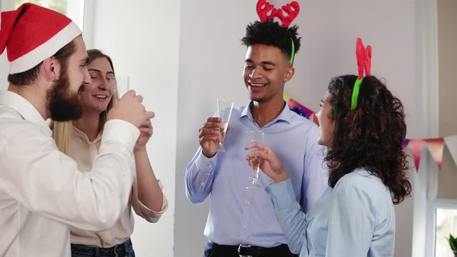 Multiethnic group of young office workers clinking glasses with sparkling wine wearing christmas hats and celebrating Christmas and New Year. Slowmotion shot