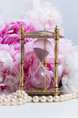 Hour glass, pearls and fresh peony flowers on white leather background
