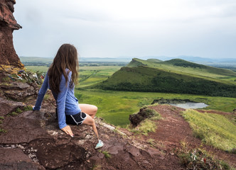 Naklejka premium The girl sits on the mountain and looks into the lake. At the bottom are green fields.