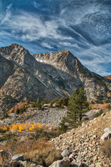 Dramatic granite faced mountains rise rapidly from a canyon with aspen trees in full fall colors. A dramatic blue sky with wispy clouds are in the background.