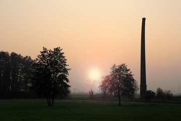 Krzywy komin zabytkowej cegielni na tle zachodzącego słońca.
