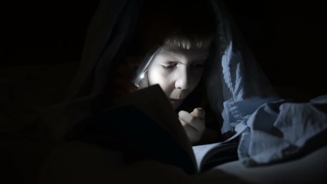A boy with a flashlight under a blanket is reading a book. Shkolnik, emotions, secrets, horrors, laughter