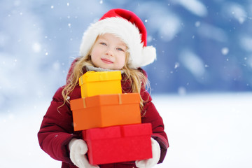 Adorable little girl wearing Santa hat holding a pile of Christmas gifts on beautiful winter day
