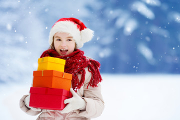 Adorable little girl wearing Santa hat holding a pile of Christmas gifts on beautiful winter day