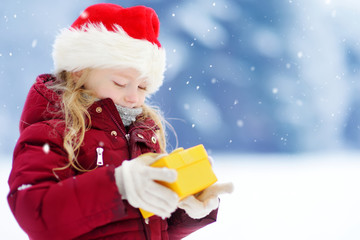 Adorable little girl wearing Santa hat holding Christmas gift on beautiful winter day
