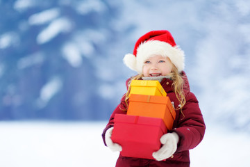 Adorable little girl wearing Santa hat holding a pile of Christmas gifts on beautiful winter day
