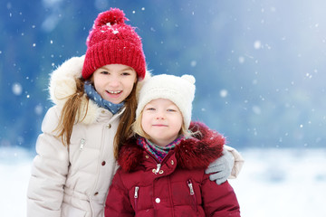 Fototapeta premium Two adorable little girls having fun together in beautiful winter park. Beautiful sisters playing in a snow.