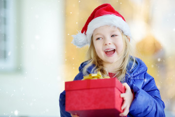 Adorable little girl wearing Santa hat holding Christmas gift on beautiful winter day