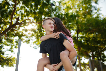 Portrait of young smiling boy holding pretty girl on his back and happily looking aside while spending time in park