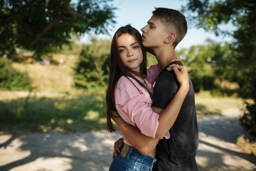 Portrait of beautiful girl thoughtfully looking in camera while standing and embracing with cool boy in park