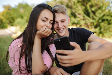 Portrait of beautiful smiling couple sitting on lawn in park and joyfully using mobile phone together