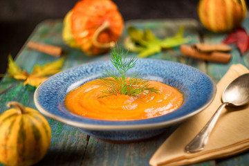 Pumpkin soup in ceramic flat dish and blue spoon. Next small decorative pumpkins and red autumn leaves. Healthy eating