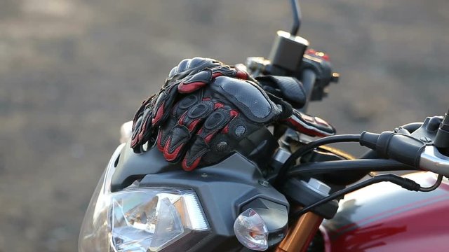 A young man in black leather jacket and white helmet sits on motorcycle puts on protective gloves before journey at autumn sunset. Close up shot.