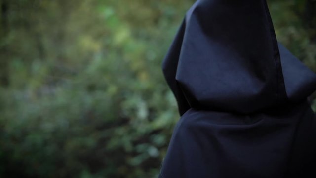 woman is wearing black raincoat with hood is walking fast in a dense scary forest in dull weather in evening