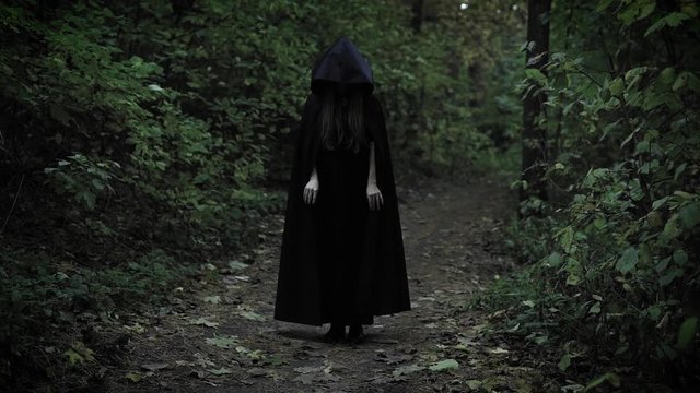 black witch is wearing black coat with hood sliding on a face, is standing still on a path in a dens dark forest