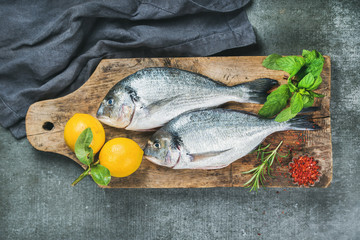 Fresh uncooked sea bream or dorado fish with lemon, herbs and spices on rustic wooden board over grey concrete background, top view. Healthy, dieting, clean eating concept