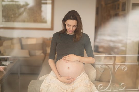 Pregnant Woman Touching Her Belly While Sitting On Sofa At Home