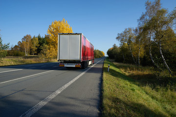 Red truck driving on the asphalt road leading through a forest in autumn colors