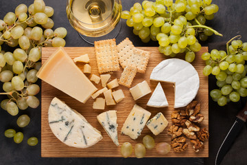 Assorted cheeses with white grapes, walnuts, crackers and white wine on a wooden Board. Food for a romantic date on a dark stone background. Top view