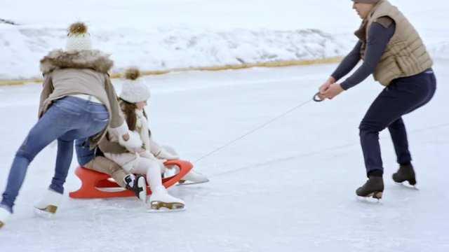 Happy young man and woman ice skating and spinning sled with laughing children on outdoor rink in winter
