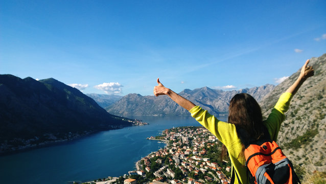 Brunette girl stands on top of a mountain and looking in the direction of Boko-Kotor Bay to Kotor in Montenegro. Traveler with backpack shows thumbs up - she's happy