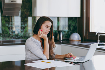 Attractive pensive and sad business woman working with documents and laptop in the kitchen at home