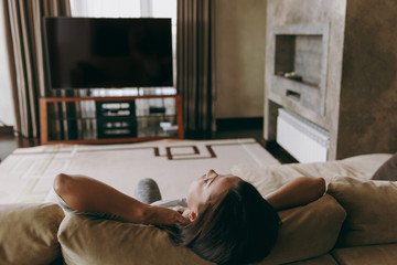 Young woman at home lying on the sofa, relaxing in her living room and watching TV. Close up. Back view. With place for text.