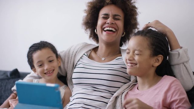 Young mother or childminder laughs with two young children as they watch a digital tablet