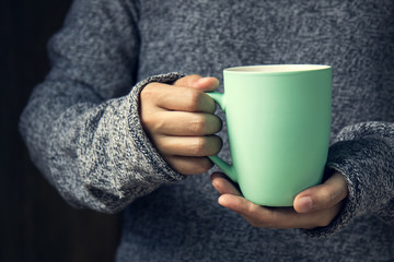 The girl in a cozy knitted sweater holding hot cup of coffee or tea in cafe or living room.