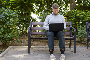 Young freelancer works on his laptop sitting on a bench in a park.