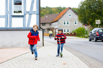 Two little schoolkids boys running and driving on scooter on autumn day