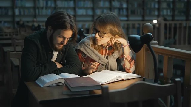 Couple of students are studying with books in the libraryhall with bookshelves, indoor dusk slowmotion