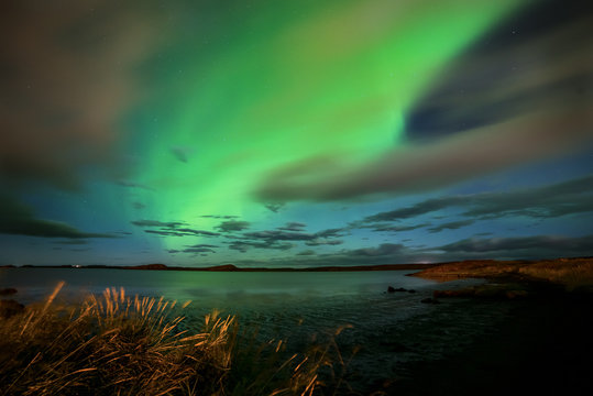 Aurora borealis on the lake shore. The radiance reflects in the water of the lake, in the background in the distance the silhouettes of the mountains. Lovely night view. Iceland.
