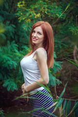 Portrait of beautiful young smiling woman with long red hair, wearing long striped skirt and white top standing in a forest