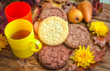 Autumn breakfast or snack. Homemade pastry – chocolate and milk flavor biscuits, tea in a colorful cups, yellow chrysanthemum flowers, dry oak leaves and plums on the table.