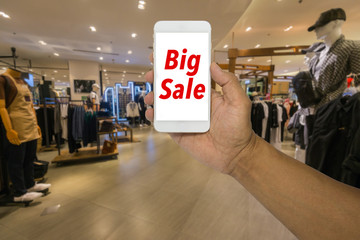 Fototapeta premium Hand holding phone in a clothes store of a shopping mall. Big sale on the screen of the smartphone