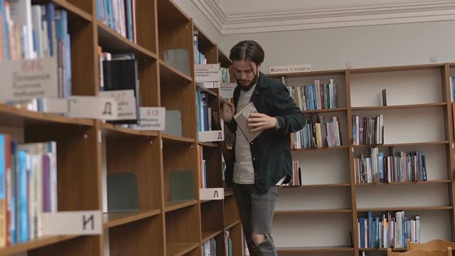 Chosing books by young long hair student man on bookshelves in big library
