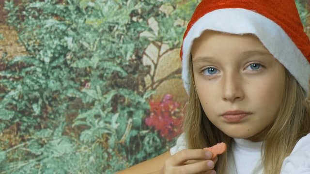 Young cute little blonde happy girl in christmas red cap eating chip ginger cookies in xmas hat and long blonde hair on garden background.