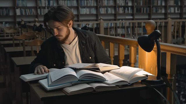 Bored student man use books for studying to exams before bookshelves in evening time in the library