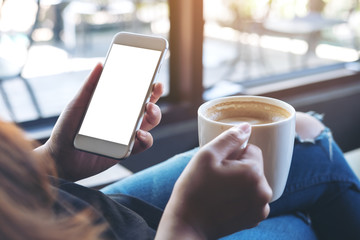 Mockup image of woman's hands holding white mobile phone with blank screen while drinking coffee in modern loft cafe
