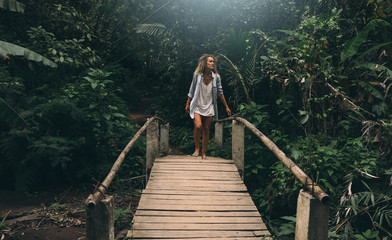 A travel blogger female is walking old wooden bridge hidden amid the wild jungles in exotic country. Young woman with curly hair is impressed with beautiful wild nature and tropical plants.