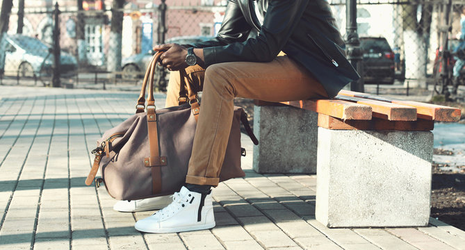 Fashion stylish man with bag sitting on a bench in the city park close up