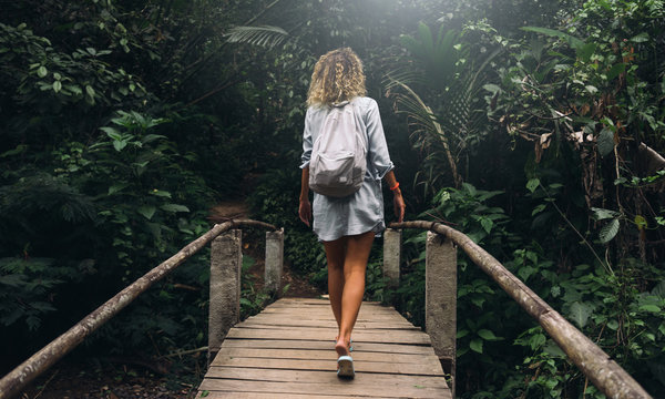 View from the back photo of young caucasian female with curly hair walking the old wooden bridge in the jungles. Backpacker female is exploring tropical forest during summer vacation.