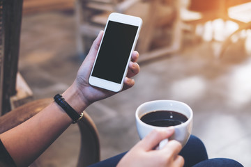 Mockup image of woman's hands holding white mobile phone with blank black screen while drinking coffee in modern loft cafe