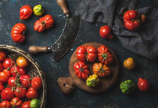 Fresh colorful ripe Fall heirloom tomatoes in basket and on wooden board, herb chopper knife for cooking over grunge dark plywood background, top view. Harvest vegetable cooking conception.