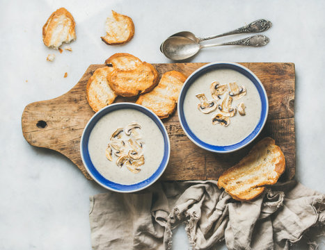 Creamy mushroom soup in bowls with toasted bread slices on rustic serving board over grey marble background, top view, selective focus