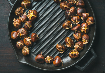 Roasted chestnuts in cast iron grilling pan over dark scorched wooden background, top view, copy space