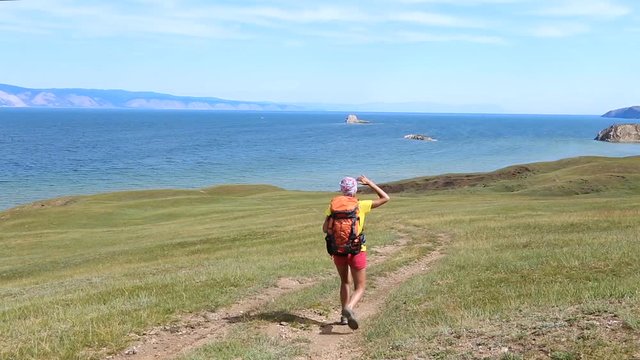 Slender hiking woman with backpack walking along the road near the blue lake Baikal.